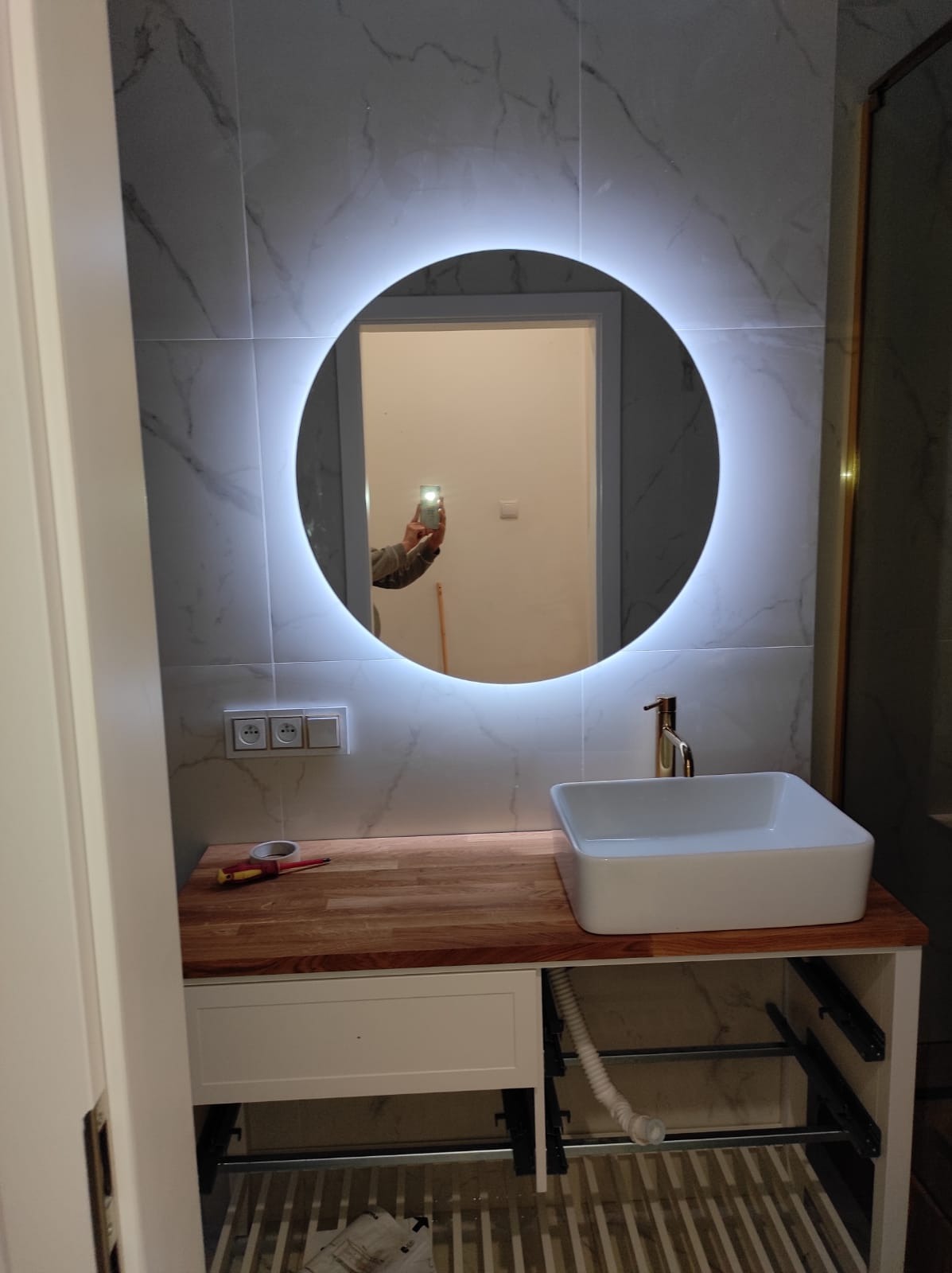 Montaż umywalki nablatowej z baterią w łazience z okrągłym lustrem podświetlanym LED, widoczne narzędzia i niedokończone podłączenie odpływu.