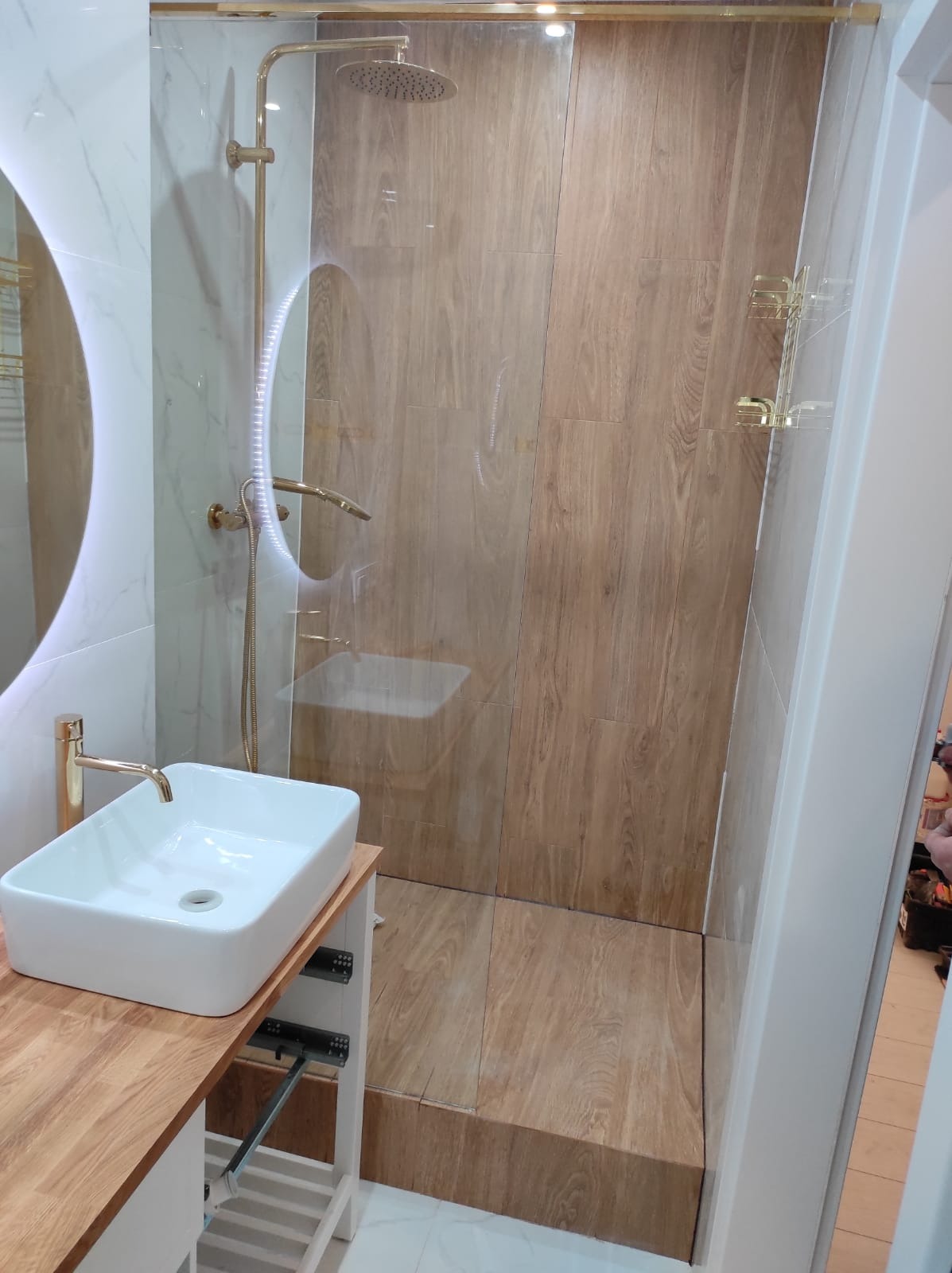 Nowoczesna łazienka z kabiną prysznicową typu walk-in wykończona płytkami imitującymi drewno, armatura w kolorze złotym, umywalka nablatowa na drewnianym blacie.