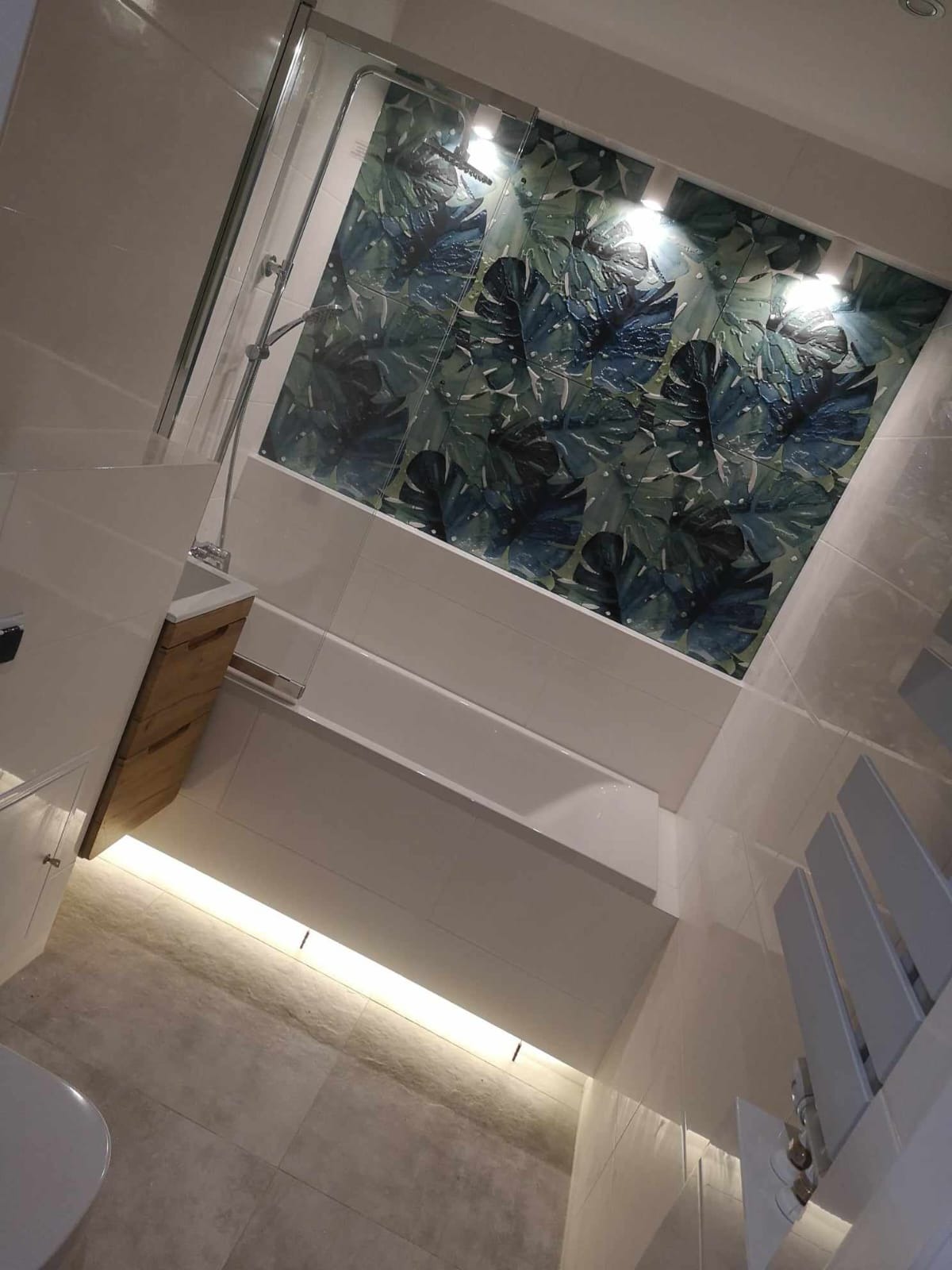 Nowoczesna łazienka z kabiną prysznicową i ścianą ozdobioną płytkami z motywem liści monstery, podświetlona listwą LED pod wanną.