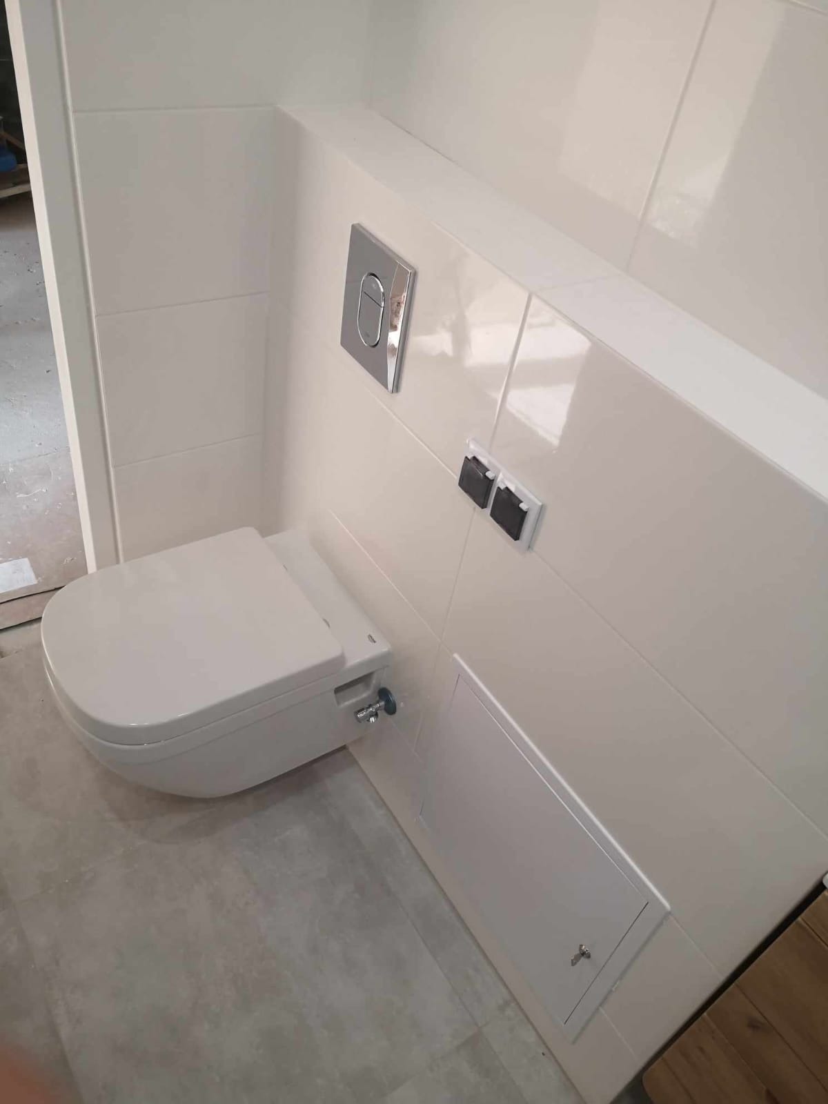 Podwieszana toaleta z funkcją bidetu, zamontowana na ścianie wyłożonej błyszczącymi, białymi płytkami; widoczny panel sterujący spłukiwaniem, szara podłoga i ukryty zawór dopływu wody.