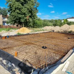 Zbrojenie fundamentów domu jednorodzinnego, widoczne pręty zbrojeniowe tworzące siatkę, rury kanalizacyjne, czarna folia izolacyjna i ocieplenie obwodowe, w tle budynek mieszkalny i drzewa.