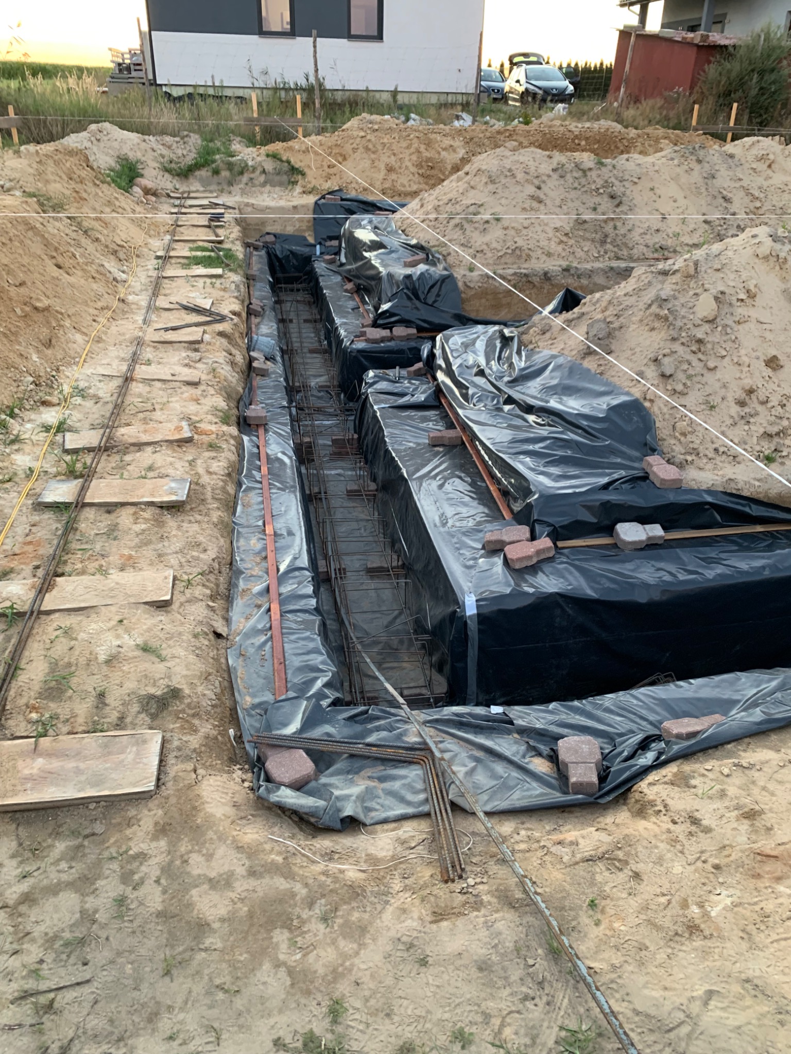 Przygotowanie wykopu pod fundamenty budynku: zbrojenie w szalunku zabezpieczonym folią, widoczne uziomy i rozłożone cegły klinkierowe, ziemia usypana po bokach.