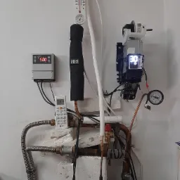 Fragment instalacji z rurami miedzianymi, zaworami, czujnikami temperatury, manometrem i sterownikiem, widoczny także pilot LG, całość zamontowana na białej ścianie.
