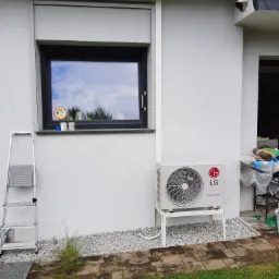 Biała jednostka zewnętrzna klimatyzacji LG Dual Inverter na metalowym stelażu, ustawiona na białym żwirze przy ścianie domu z oknem z roletą i drabinką opartą o ścianę.