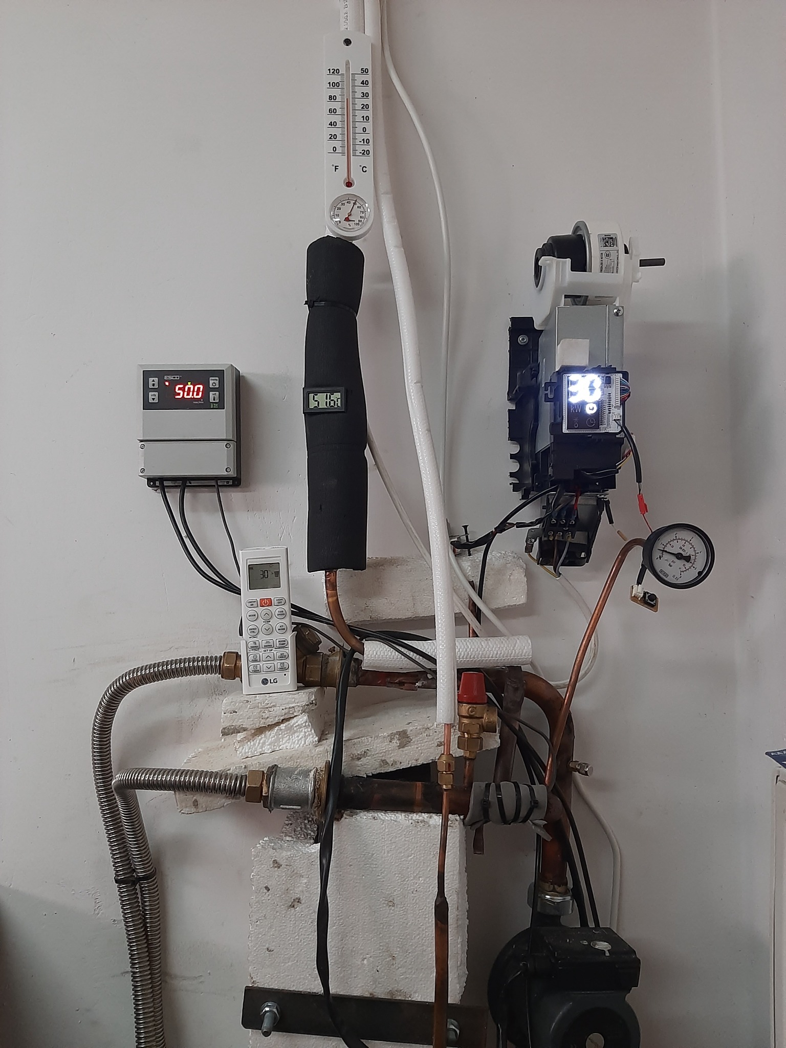 Fragment instalacji z rurami miedzianymi, zaworami, czujnikami temperatury, manometrem i sterownikiem, widoczny także pilot LG, całość zamontowana na białej ścianie.