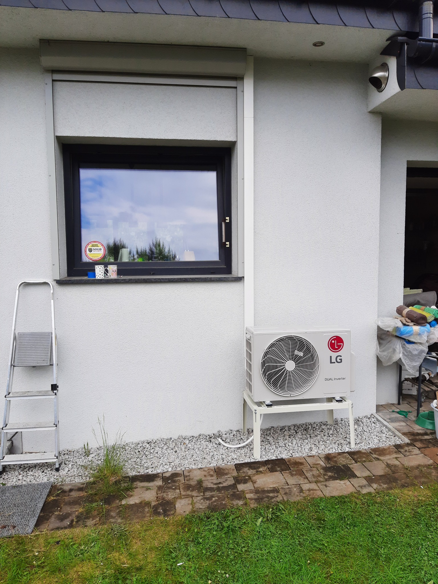 Biała jednostka zewnętrzna klimatyzacji LG Dual Inverter na metalowym stelażu, ustawiona na białym żwirze przy ścianie domu z oknem z roletą i drabinką opartą o ścianę.