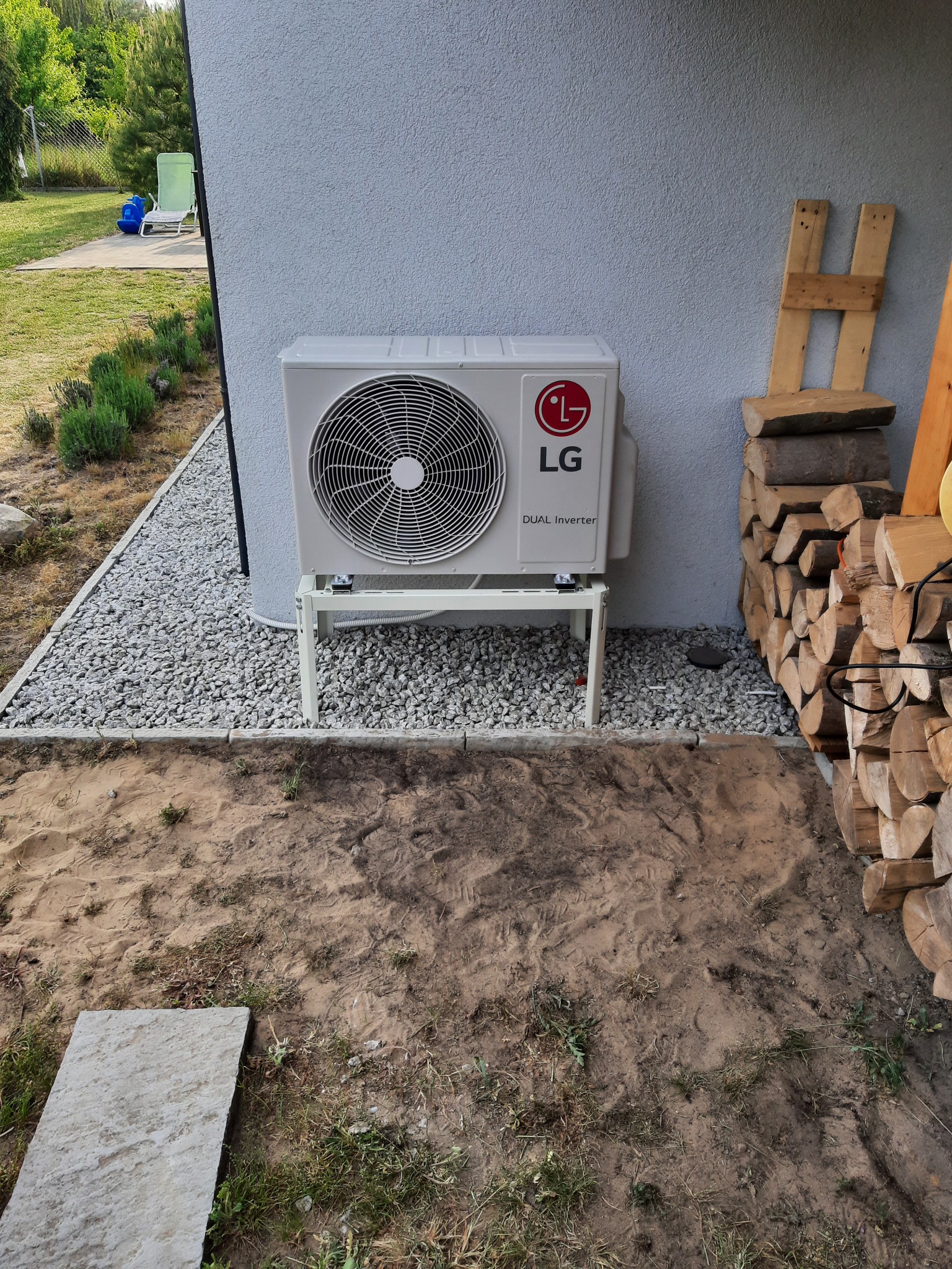 Zewnętrzna jednostka klimatyzacji LG Dual Inverter zamontowana na białym stelażu przy ścianie domu, obok ułożone drewno kominkowe, widoczny fragment trawnika i krzewów lawendy.