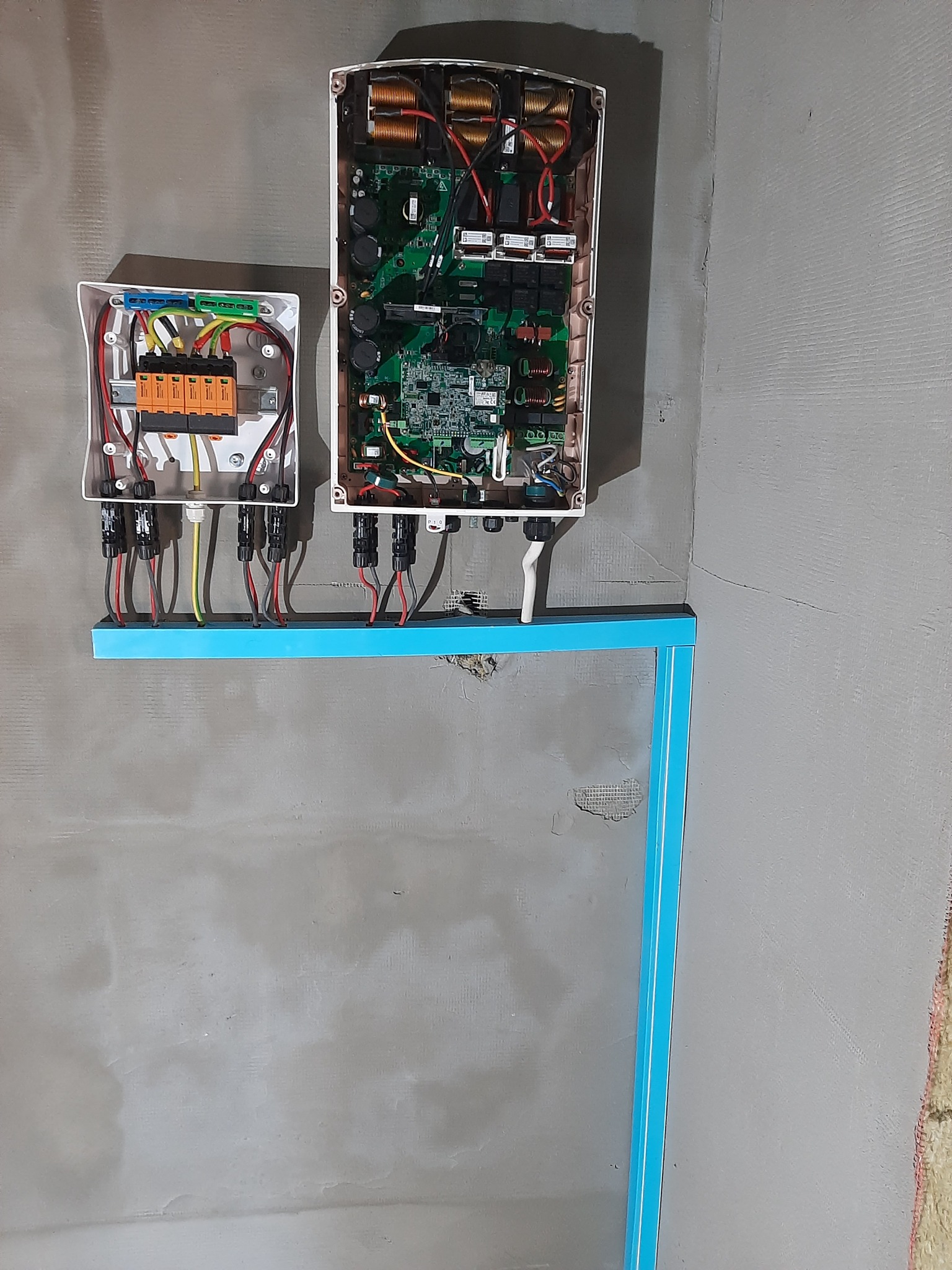 Dwa otwarte panele elektryczne z widocznymi obwodami i kablami, zamontowane na niebieskim wsporniku na szarej, surowej ścianie.