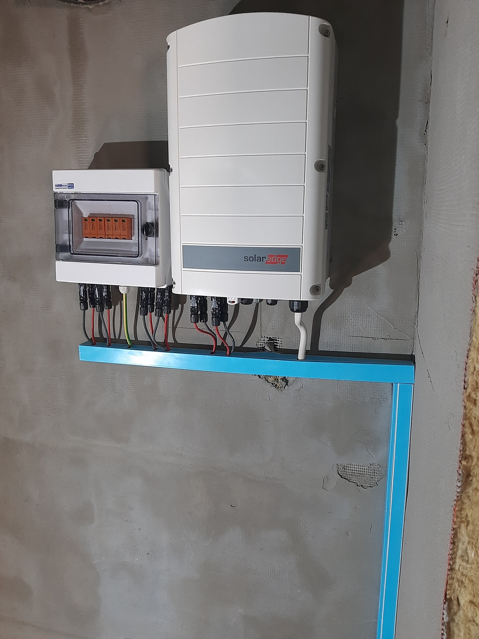 Biała obudowa falownika SolarEdge zamontowana na surowej, szarej ścianie wraz z podłączeniami elektrycznymi i niebieskim profilem mocującym.