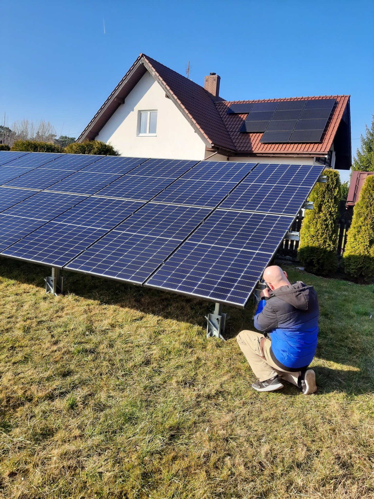 Instalator w trakcie montażu paneli słonecznych na trawniku przed domem z panelami fotowoltaicznymi na dachu, widoczne narzędzia i elementy konstrukcyjne.