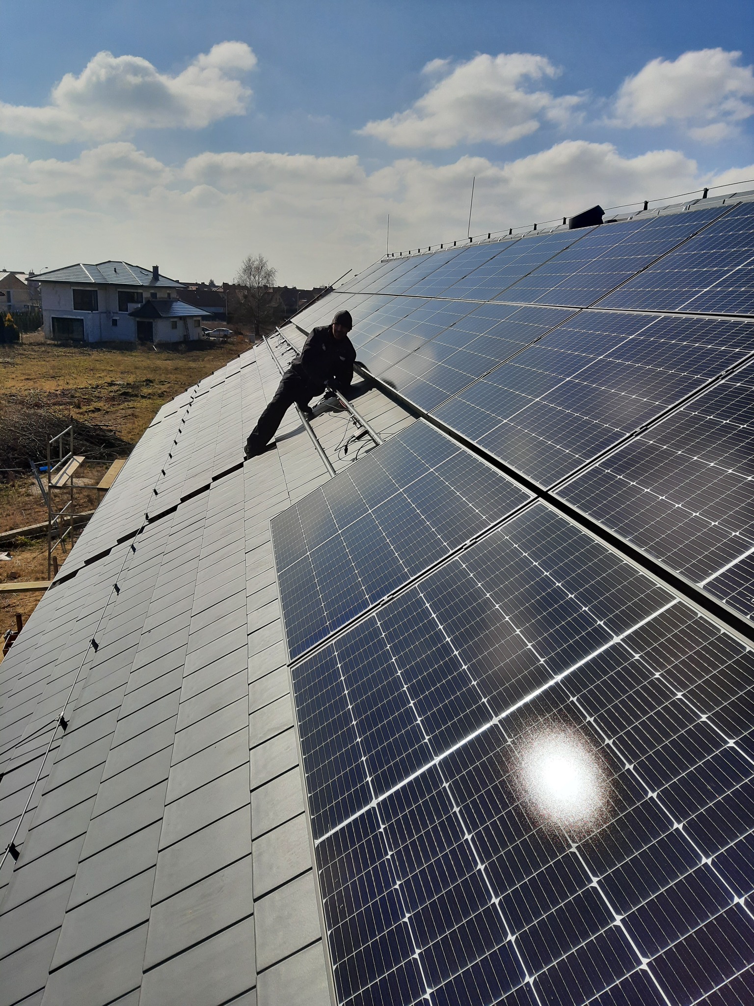 Montaż paneli słonecznych na dachu pokrytym szarą dachówką; instalator na drabinie, odbicie słońca na panelach, dom w tle.