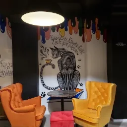 Wnętrze kawiarni z muralem przedstawiającym kota pijącego kawę, pomalowane ściany w stylu drip art, dwa fotele w kolorach pomarańczowym i żółtym oraz czerwony puf.