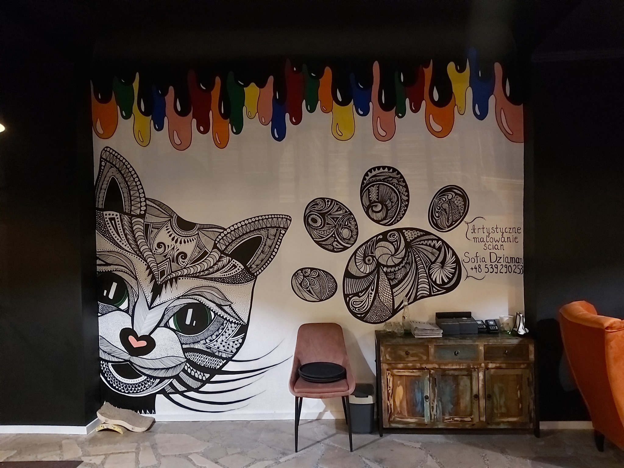Ściana z artystycznym malunkiem przedstawiającym głowę kota i odciski łap w stylu mandali, nad którymi widnieją kolorowe krople farby, obok stoi różowe krzesło i rustykalna komoda.