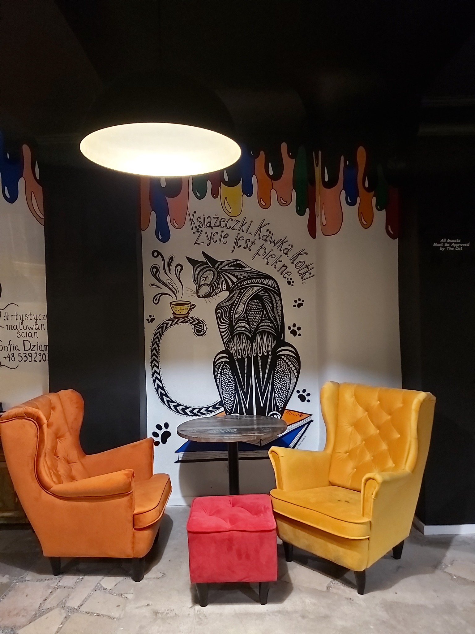 Wnętrze kawiarni z muralem przedstawiającym kota pijącego kawę, pomalowane ściany w stylu drip art, dwa fotele w kolorach pomarańczowym i żółtym oraz czerwony puf.