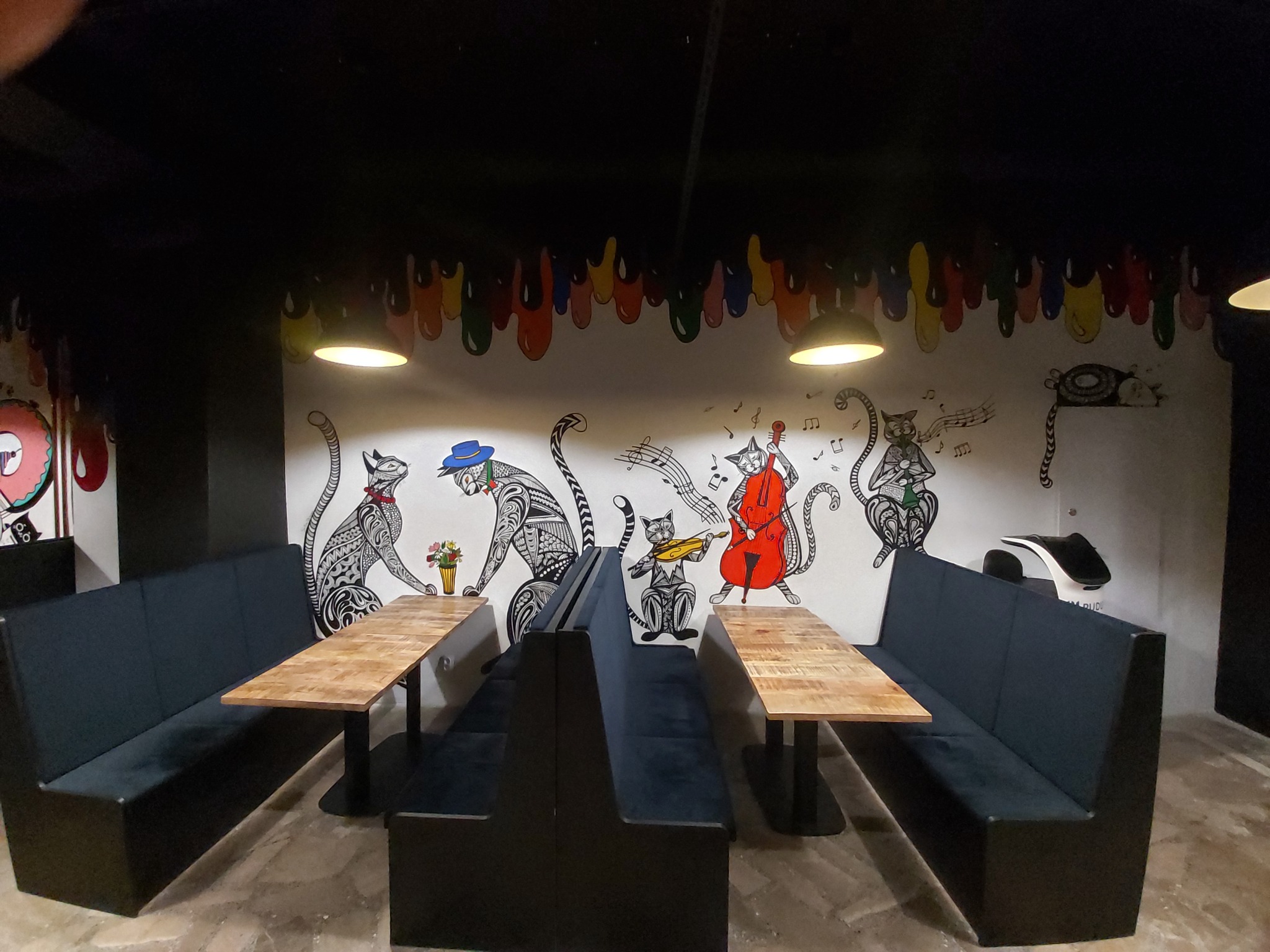 Wnętrze lokalu gastronomicznego z muralem przedstawiającym koty grające na instrumentach, drewniane stoły i ciemne sofy w boksach.