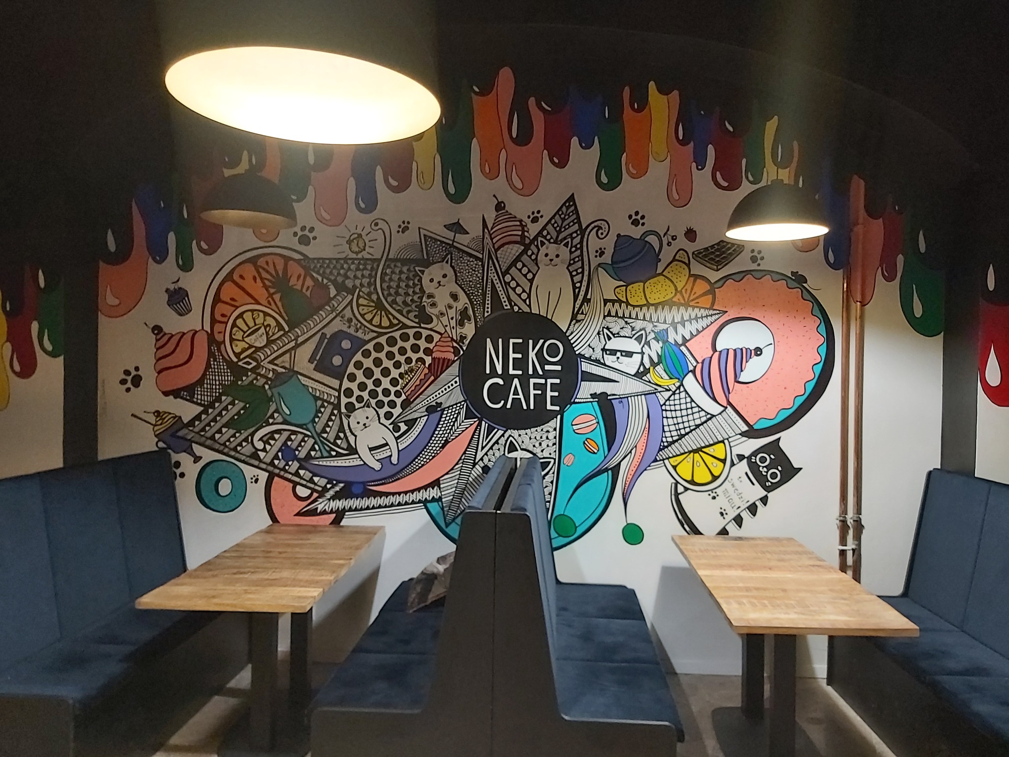 Wnętrze kawiarni z muralem przedstawiającym koty, słodycze i napisy w stylu graffiti, oświetlone dwoma lampami sufitowymi, z drewnianymi stolikami i tapicerowanymi ławami.