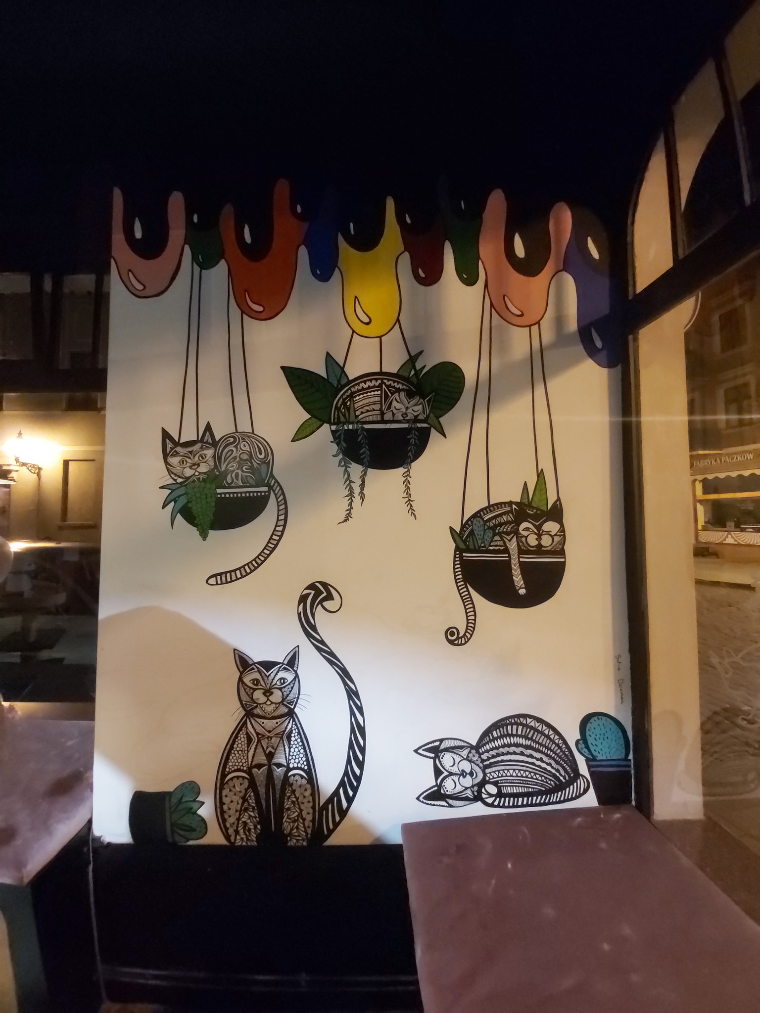 Ilustracja na ścianie przedstawiająca koty w doniczkach wiszących pod kolorowymi plamami farby, wykonana w stylu linearnym, z widocznymi elementami roślinnymi i geometrycznymi wzorami.