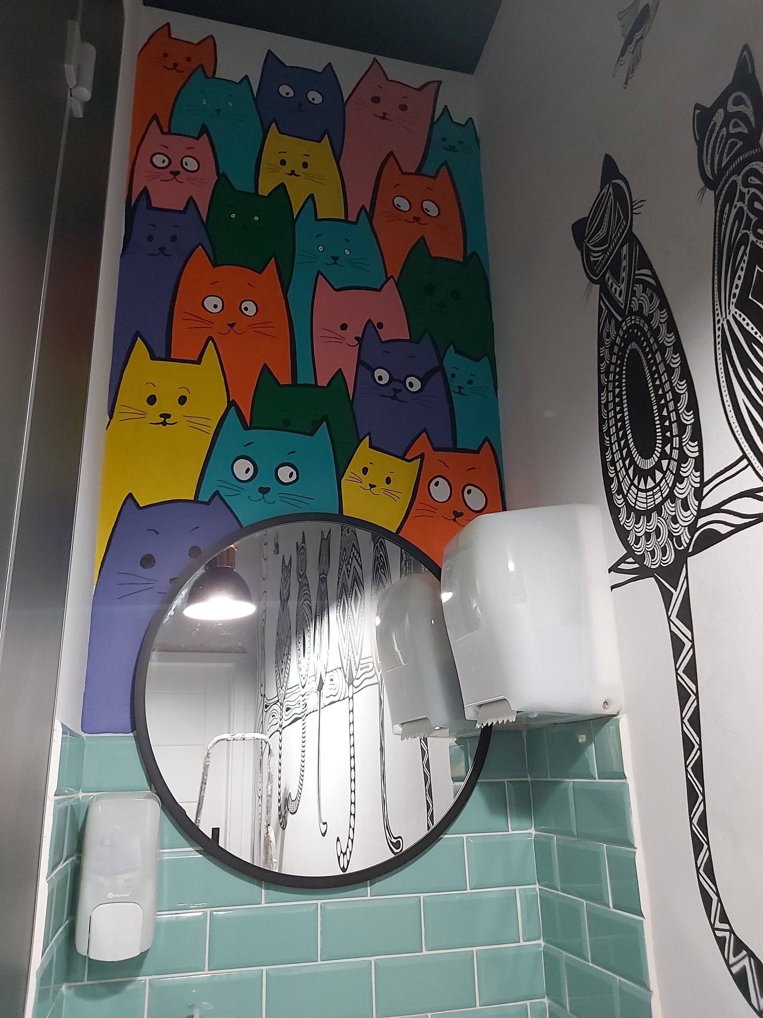 Kolorowa ściana z rysunkami kotów w łazience, odbicie w okrągłym lustrze, turkusowe płytki, podajnik na ręczniki.