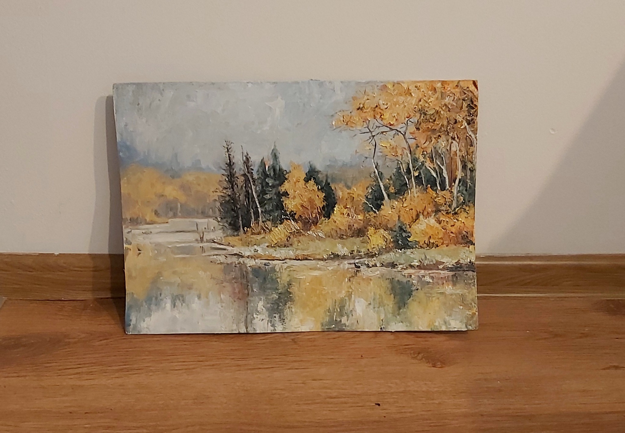 Jesienny pejzaż z rzeką namalowany farbami olejnymi, przedstawiający odbicie drzew w wodzie, wykonany przez artystę z Piły. Obraz leży na drewnianej podłodze.