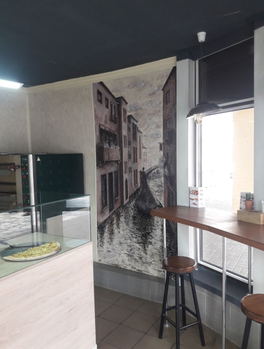 Wnętrze lokalu z muralem przedstawiającym wenecki pejzaż, z pizzą w witrynie i wysokimi stołkami barowymi przy oknie.