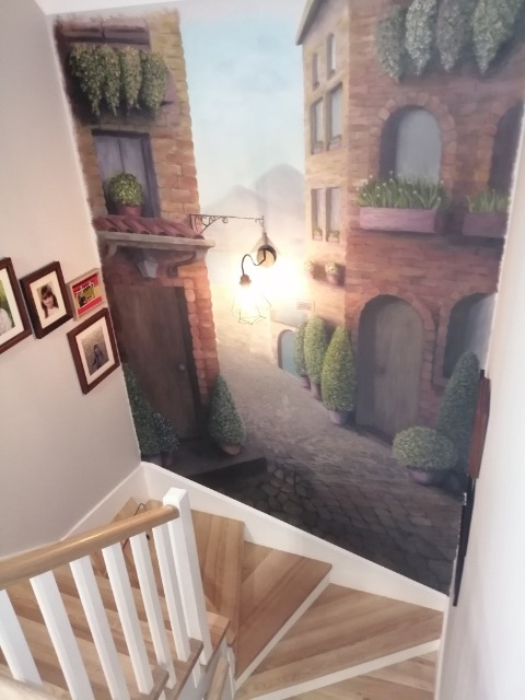 Mural na ścianie klatki schodowej przedstawiający uliczkę włoskiego miasteczka z brukowaną drogą, budynkami z cegły, lampionem i roślinami w donicach, widziany z góry schodów z drewnianymi...