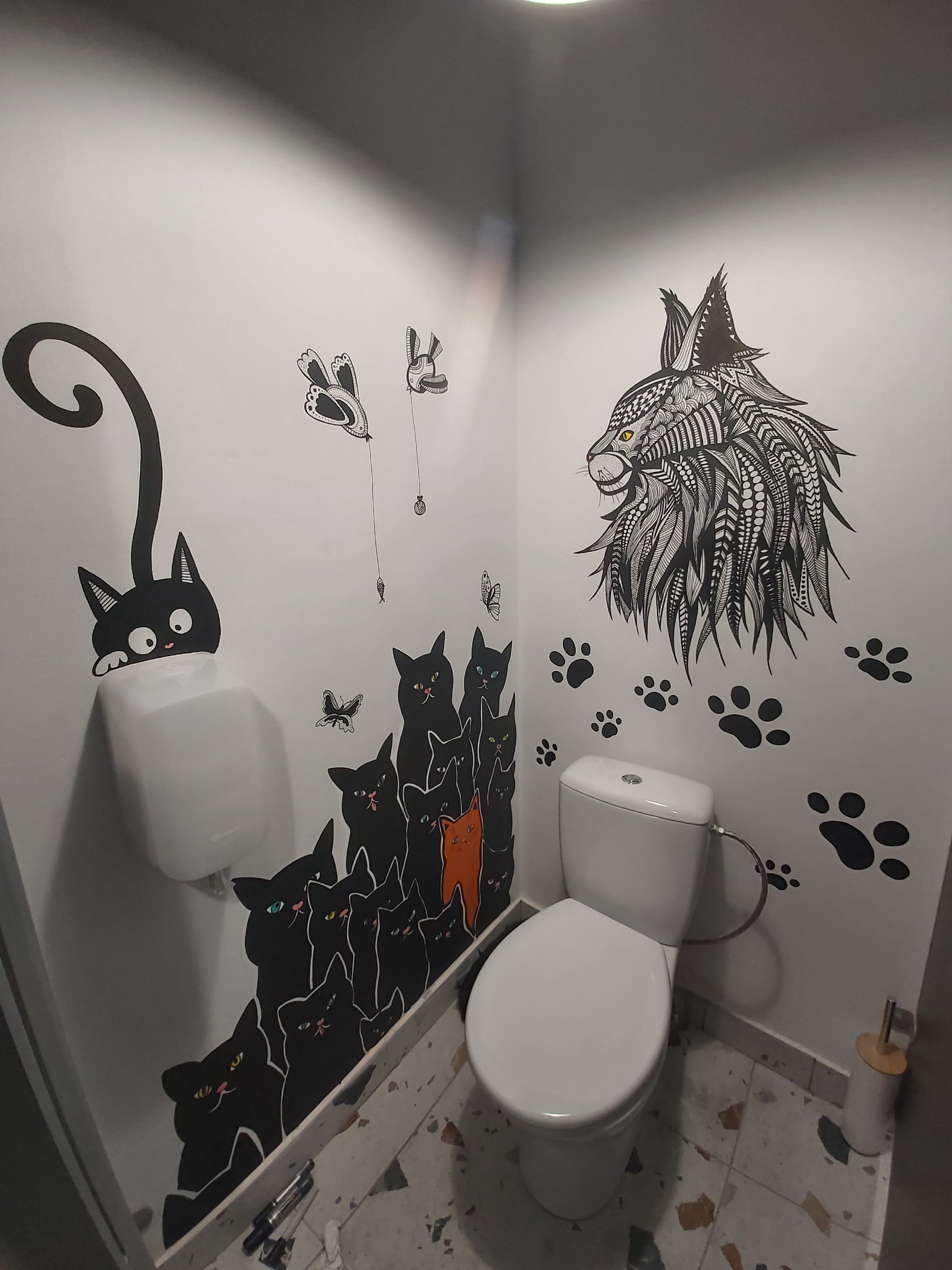 Wnętrze toalety z artystycznym malowaniem ścian przedstawiającym czarne koty, w tym jeden spoglądający znad pisuaru, oraz rysunek rysia wykonany w stylu doodle art. Na ścianach widoczne także...