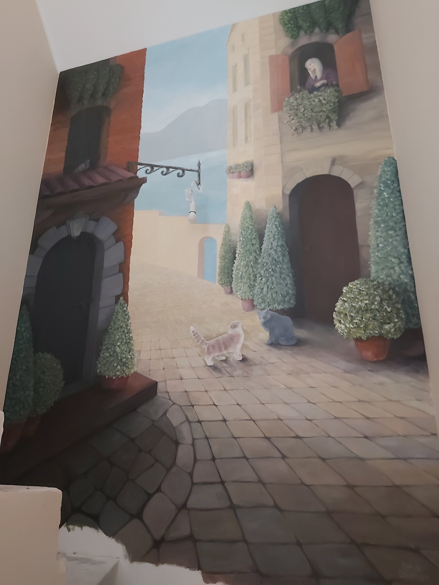 Iluzjonistyczne malowidło ścienne przedstawiające włoską uliczkę z dwoma kotami, drzewami w donicach i postacią w oknie, wykonane w ciepłej tonacji z wyraźnym efektem perspektywy.