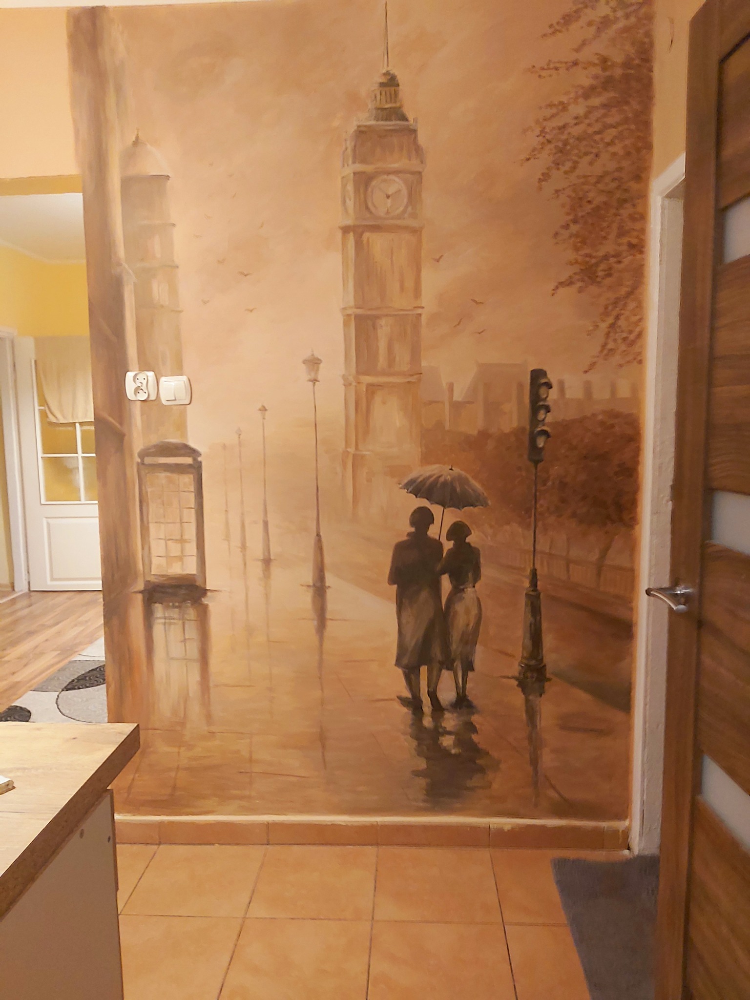 Iluzjonistyczne malowidło ścienne przedstawiające Londyn w sepii: Big Ben, budka telefoniczna, para pod parasolem, mokra ulica odbijająca światła.