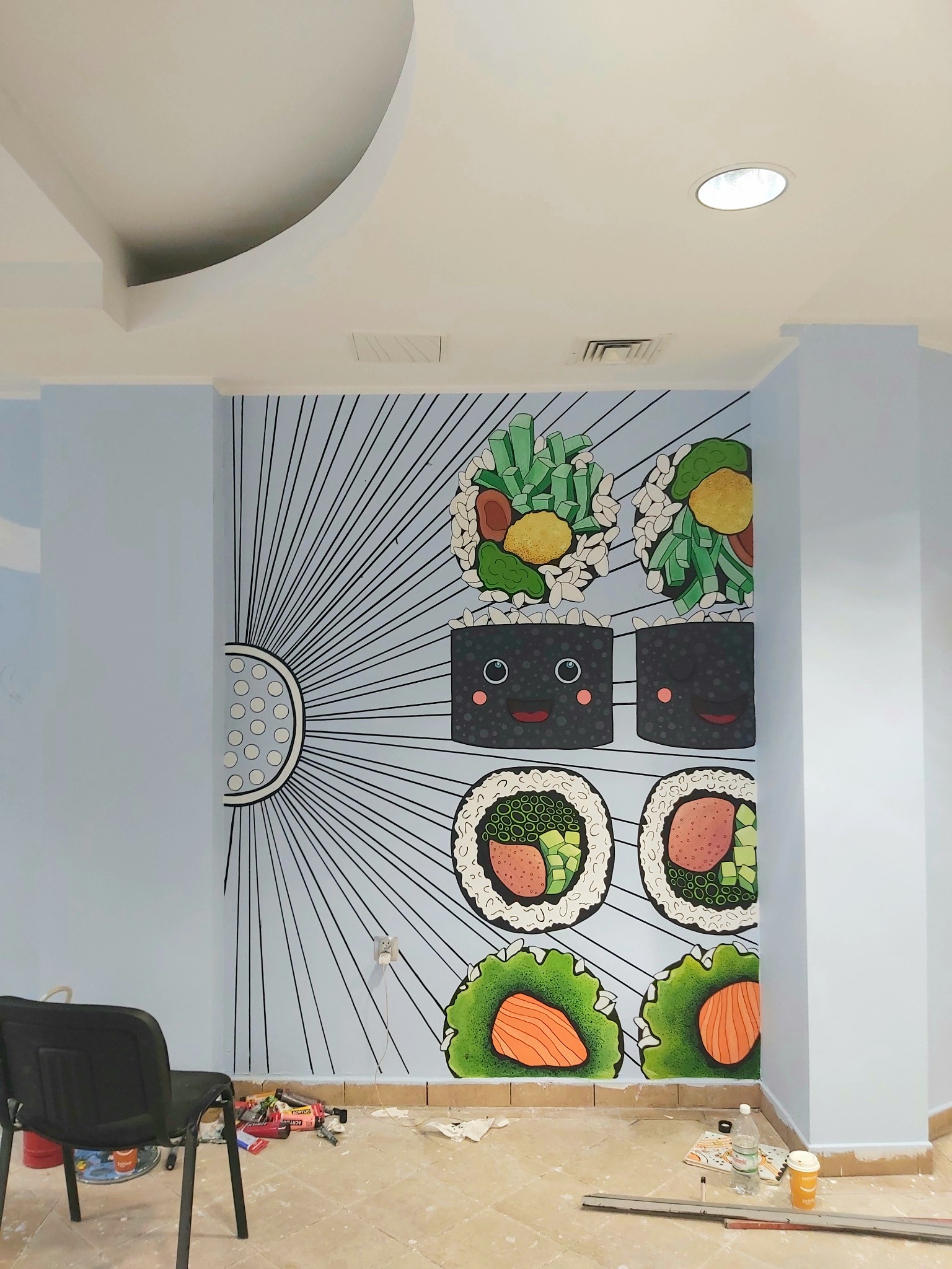Ściana ozdobiona ręcznie malowanym muralem przedstawiającym stylizowane sushi, widoczne narzędzia malarskie i krzesło w pomieszczeniu w trakcie prac wykończeniowych.