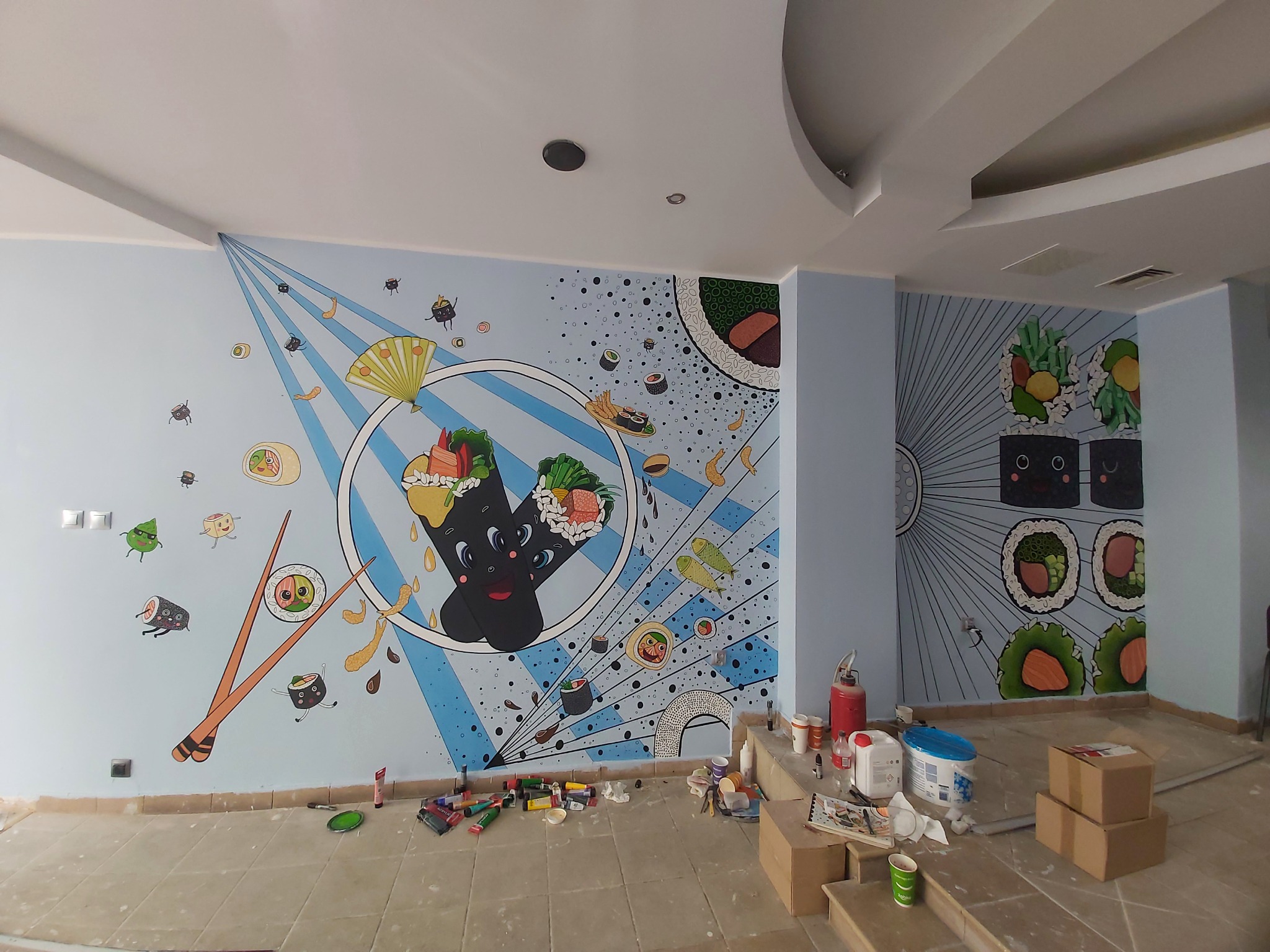 Mural z motywem sushi w stylu kawaii na jasnoniebieskiej ścianie, z widocznymi farbami i narzędziami malarskimi na podłodze w trakcie realizacji.