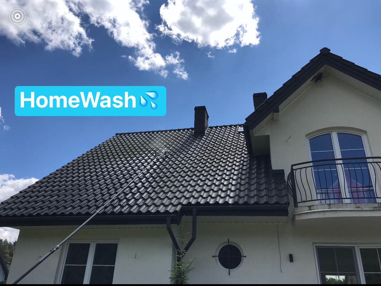 Dwupiętrowy dom z czarnym dachem z dachówki podczas czyszczenia z użyciem lancy teleskopowej. Widoczny napis 'HomeWash' z symbolem kropli wody.