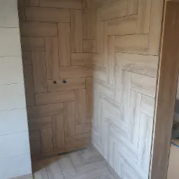 Ukończona kabina prysznicowa z drewnopodobnymi płytkami ułożonymi w jodełkę, widoczny odpływ liniowy i przygotowane przyłącza wodne.