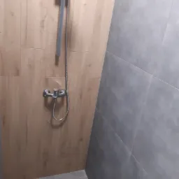 Nowoczesna kabina prysznicowa z drewnopodobnymi płytkami na ścianie, szara glazura na sąsiedniej ścianie i odpływ liniowy w podłodze.