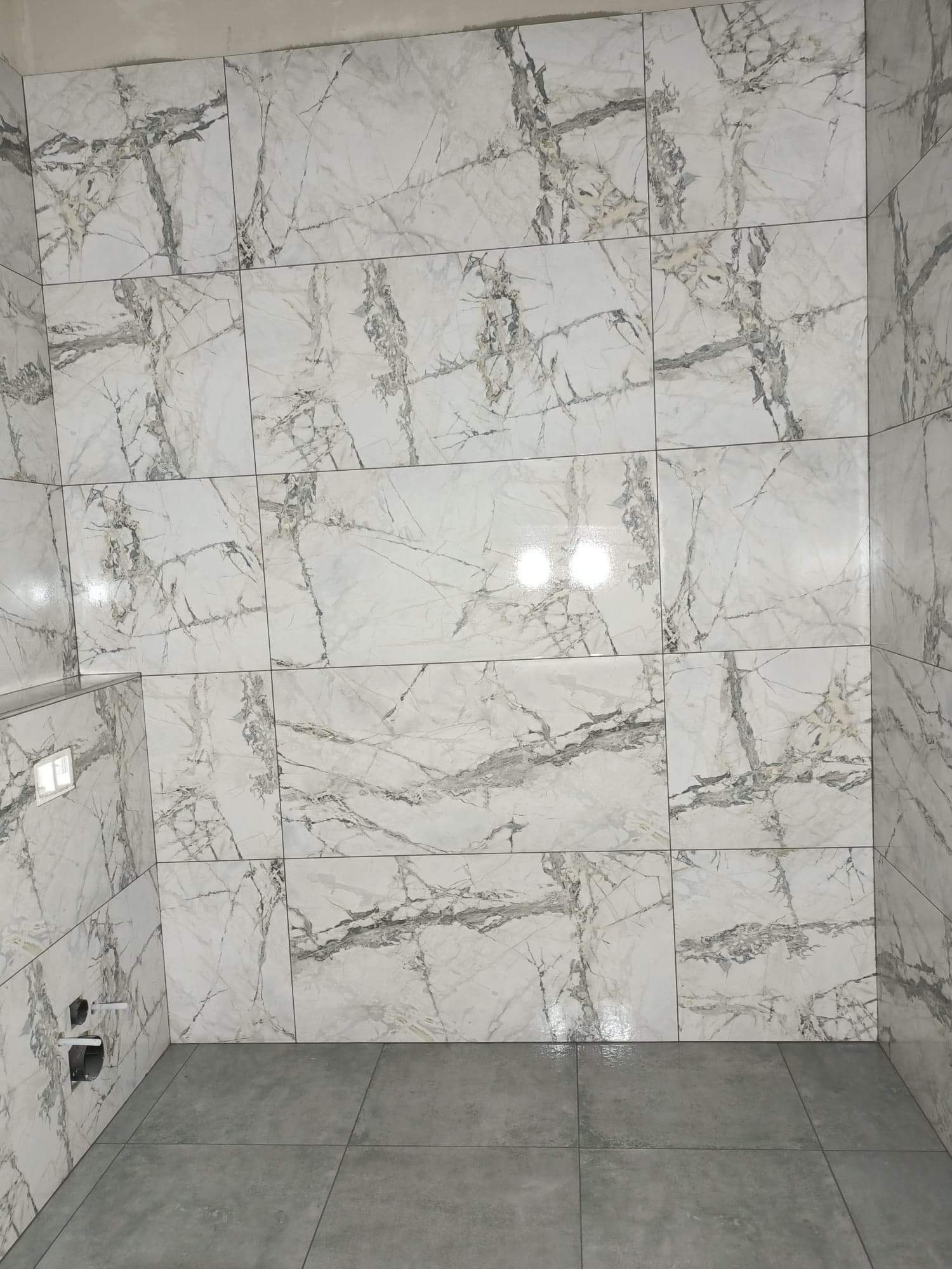 Wykończona łazienka z marmurowymi płytkami na ścianach i szarymi płytkami na podłodze, widoczne przyłącza wodno-kanalizacyjne w rogu.