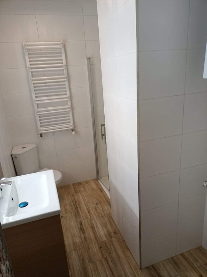 Jasna łazienka z drewnopodobną podłogą, białymi płytkami ściennymi, grzejnikiem drabinkowym, umywalką z szafką w kolorze drewna i kabiną prysznicową.