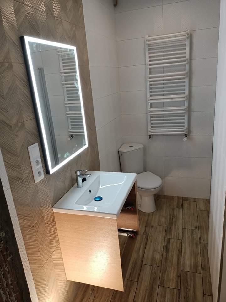 Nowoczesna łazienka z umywalką nablatową na szafce z jasnego drewna, podświetlanym lustrem i grzejnikiem drabinkowym na białych płytkach.