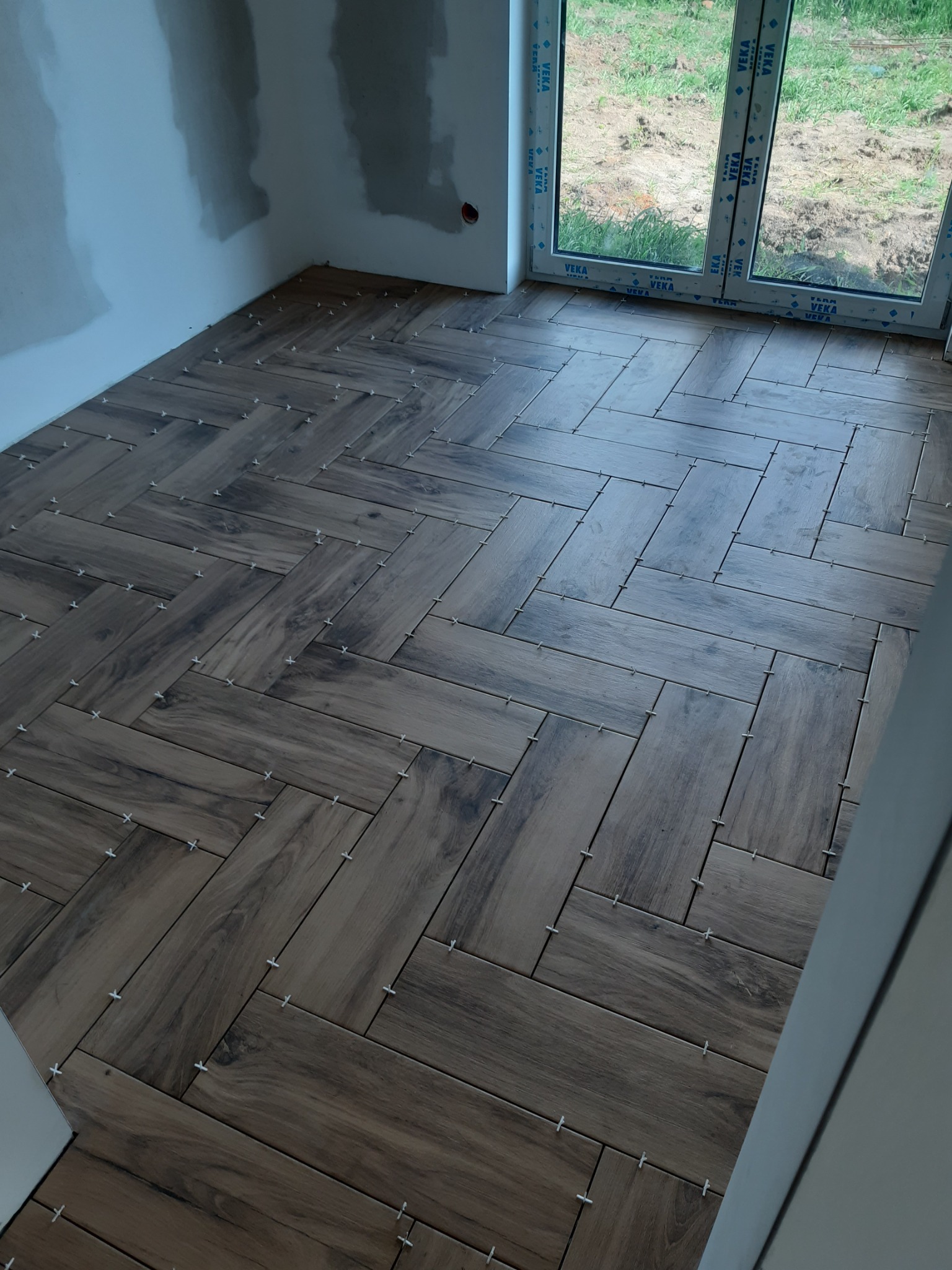Ułożona w jodełkę podłoga z płytek imitujących drewno, z białymi krzyżykami dystansowymi, widok na okno i surowe ściany w trakcie prac wykończeniowych.