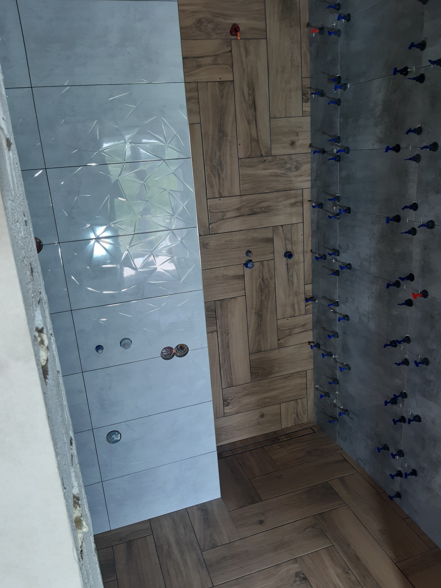 Układanie płytek: Ściana z szarych płytek z niebieskimi klipsami poziomującymi, obok ściana z białych płytek z wzorem geometrycznym i drewnopodobna podłoga ułożona w jodełkę.