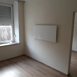 Wnętrze pokoju z ułożonymi panelami podłogowymi, białym grzejnikiem na ścianie i oknem z żaluzjami, widoczne ślady po niedawnych pracach wykończeniowych.