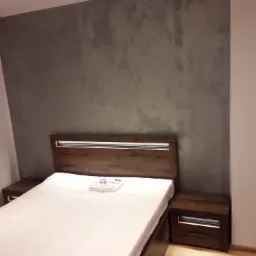 Sypialnia z łóżkiem z drewnianym wezgłowiem i szafkami nocnymi, ściana pomalowana farbą strukturalną w odcieniach szarości, pościel w folii na łóżku.