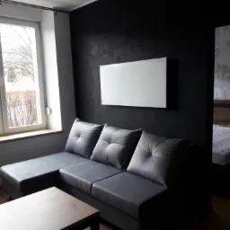 Nowoczesny salon z szarą sofą narożną, stolikiem kawowym i czarną ścianą z białym panelem grzewczym, widok na sypialnię w tle.