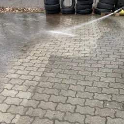 Strumień wody z węża pod ciśnieniem spłukuje brud z betonowej kostki brukowej, obok stos opon i zaparkowany biały bus.