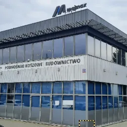 Dwukondygnacyjny budynek biurowo-magazynowy w szaro-białej kolorystyce z dużymi oknami, z widocznym logo firmy 'Majmax' na dachu i napisem 'Podnośniki Koszowe Budownictwo' nad wejściem.