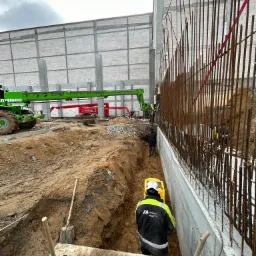 Budowa hali: robotnik w wykopie obsługuje żółty niwelator laserowy, widoczne zbrojenia fundamentów, w tle podnośniki koszowe i surowa ściana z bloczków.