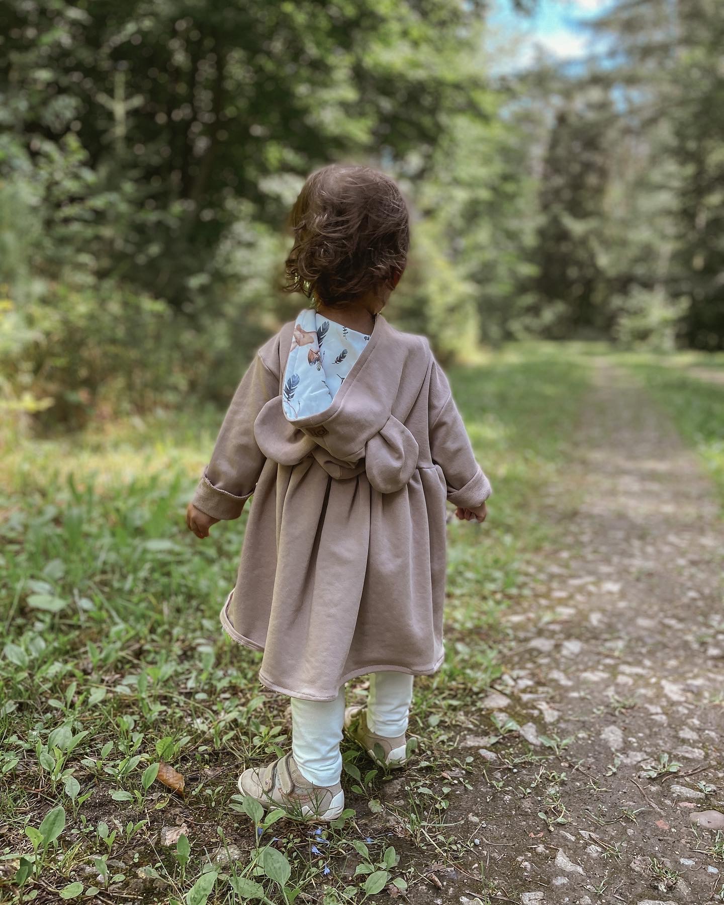 Dziewczynka w beżowej sukience z kapturem z motywem leśnym, spacerująca leśną ścieżką, widok z tyłu.