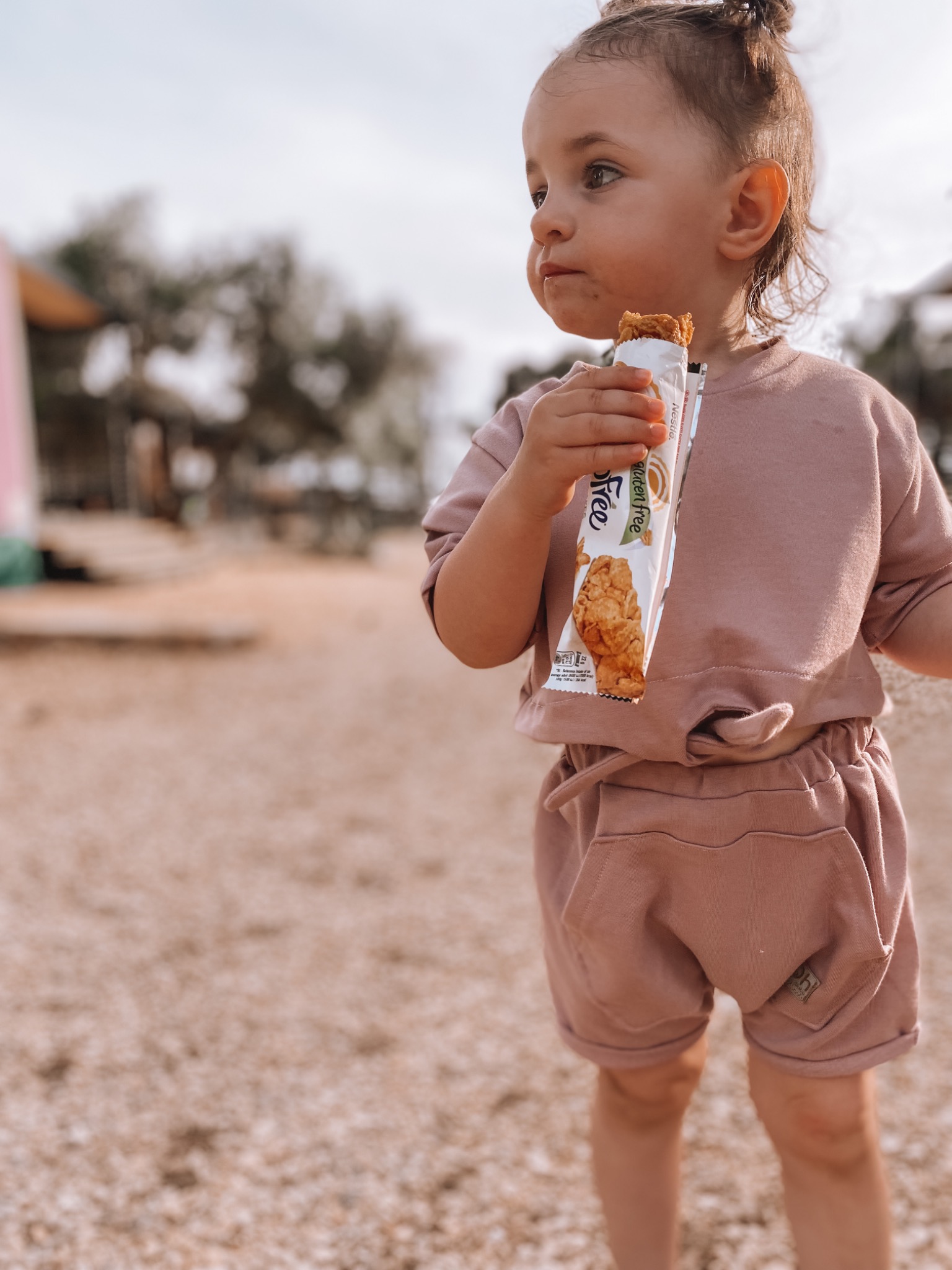 Dziewczynka w komplecie dresowym w kolorze pudrowego różu, z kieszeniami na szortach, trzymająca batonik zbożowy na tle piaszczystego placu zabaw.