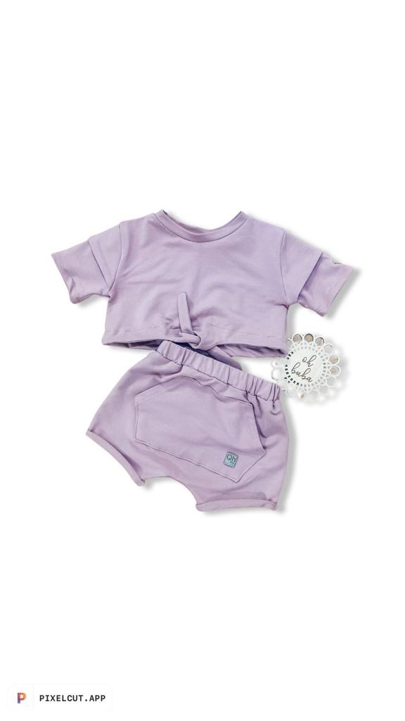 Komplet dziecięcy: krótka bluzka z wiązaniem i szorty z kieszenią, w pastelowym fiolecie, na białym tle, obok ozdobny, ażurowy kartonik z napisem.