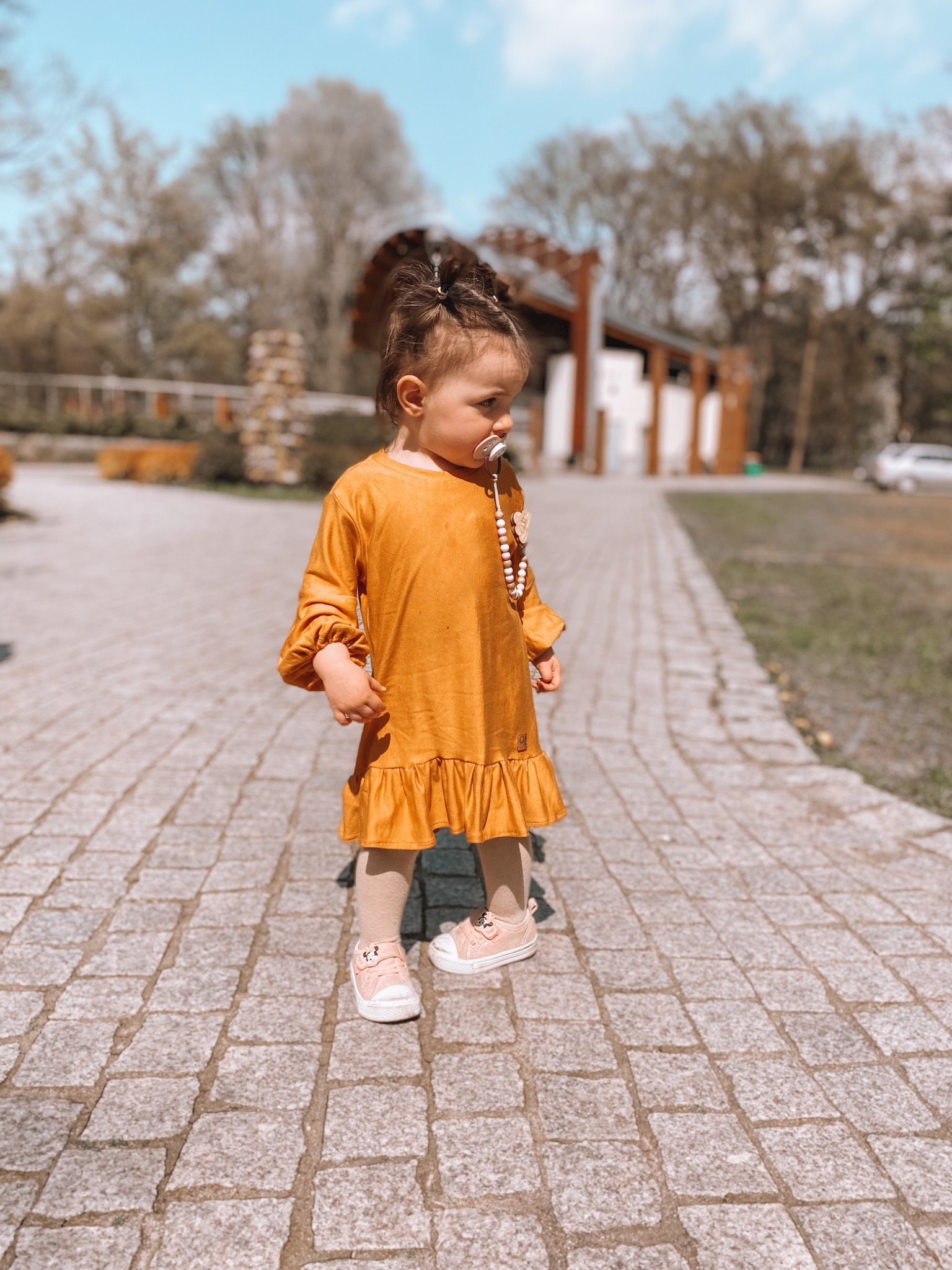 Dziewczynka w musztardowej sukience z falbanką i różowych bucikach, z smoczkiem na szyi, spaceruje po kamiennej ścieżce w słoneczny dzień.