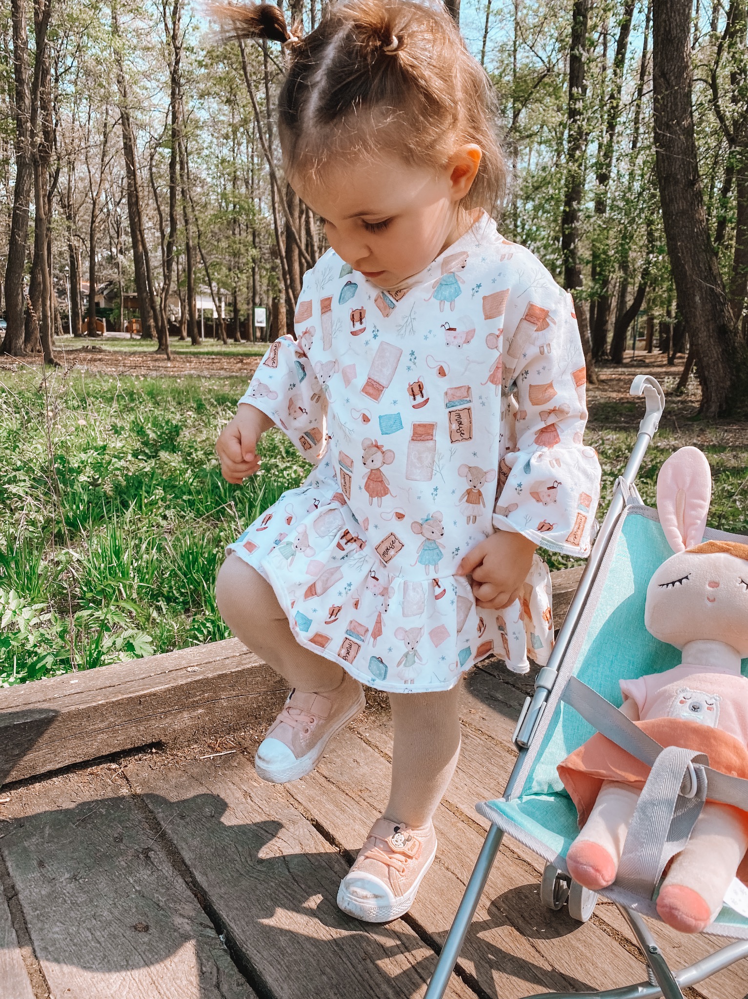 Dziewczynka w sukience z motywem myszek i książek, w różowych bucikach, stoi na drewnianym pomoście w parku, obok wózeczka z pluszowym królikiem.