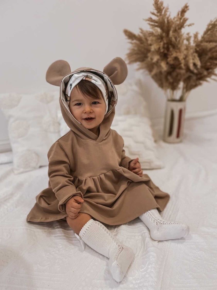 Uśmiechnięta dziewczynka w beżowej sukience z kapturem z uszami siedzi na łóżku w Warszawie. W tle wazon z suszonymi trawami.